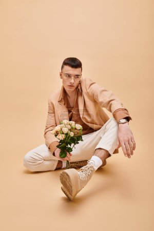 Téléchargez les photos : Beau homme en veste beige et lunettes assises avec des roses sur fond beige pêche - en image libre de droit
