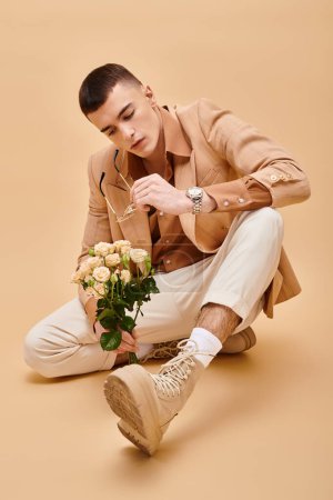 Téléchargez les photos : Prise de vue mode de l'homme en veste beige assis avec des roses et des lunettes sur fond beige pêche - en image libre de droit