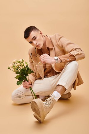 Photo for Handsome man in beige jacket sitting with roses on peachy beige background looking at camera - Royalty Free Image