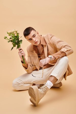Stylish man in beige jacket sitting with roses and glasses on beige background looking at camera