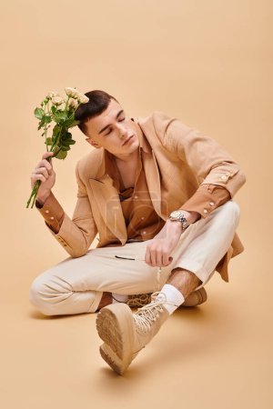 Handsome man in beige jacket sitting with roses and glasses on beige background looking away