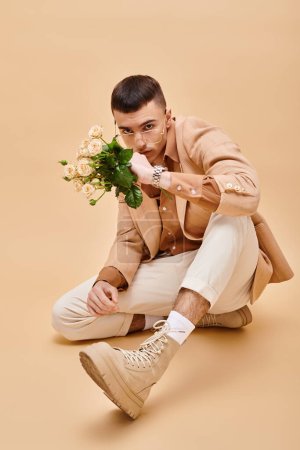 Téléchargez les photos : Jeune homme en veste beige assis avec des roses et des lunettes sur fond beige regardant la caméra - en image libre de droit