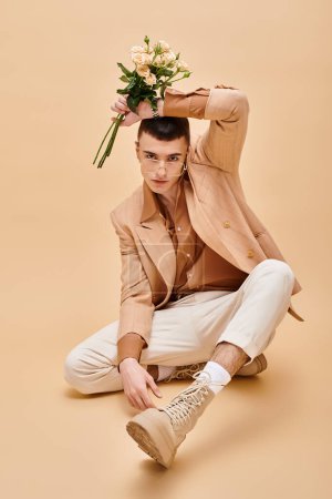 Téléchargez les photos : Homme élégant en veste beige assis avec des fleurs et des lunettes sur fond beige regardant la caméra - en image libre de droit