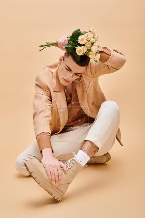 Fashionable man in beige jacket sitting with flowers and glasses on beige background looking down