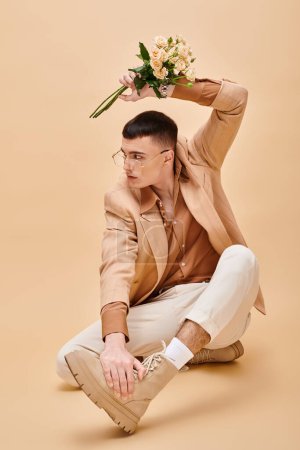 Handsome man in beige jacket sitting with roses and glasses on beige background looking away