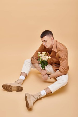 Fashionable man in beige shirt sitting with flowers and glasses on beige background looking down