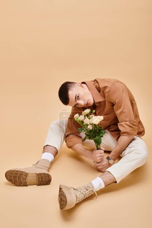 Young man in beige shirt sitting with flowers and glasses on beige background looking at camera