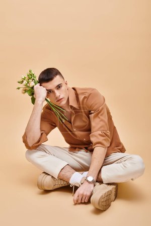 Téléchargez les photos : Portrait de mode d'homme en chemise beige assis avec des fleurs roses sur fond beige - en image libre de droit