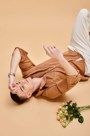 Téléchargez les photos : Portrait de mode d'homme en chemise beige couché avec bouquet de fleurs de rose sur fond beige - en image libre de droit