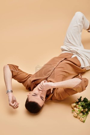 Fashion image of man in beige shirt lying with rose flowers bouquet on beige background