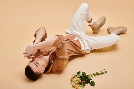 Portrait of handsome man in beige shirt lying with rose flowers bouquet on beige background