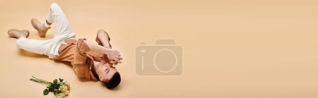 Téléchargez les photos : Portrait de bel homme en chemise beige couché avec bouquet de fleurs de rose sur fond beige, bannière - en image libre de droit