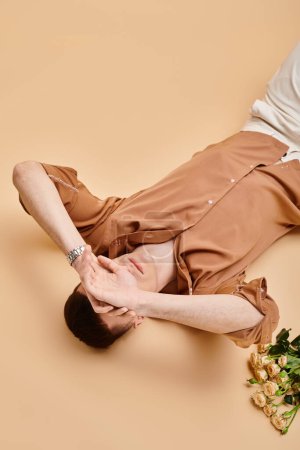 Portrait of man covering eyes with hands in beige shirt lying with rose flowers on beige background