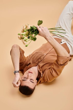 Fashion image of man in beige shirt lying with rose flowers bouquet on beige background