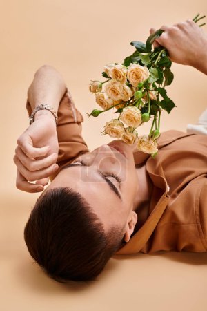 Portrait of handsome man in beige shirt lying with rose flowers bouquet on beige background