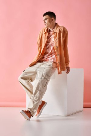 Foto de Fashionable man in beige shirt, pants and boots sitting on white cube on pink background - Imagen libre de derechos