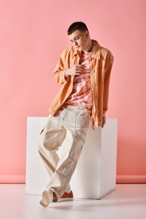 Fashion shot of fashionable man in beige shirt, pants and boots on white cube on pink background