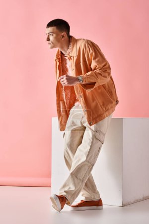 Full length image of stylish man in beige shirt, pants and boots on white cube on pink background