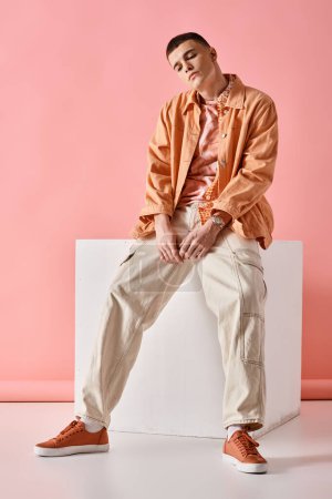 Handsome man in beige shirt, pants and sneakers sitting on white cube on pink background