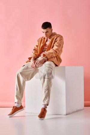 Stylish man in beige shirt, pants and sneakers sitting on white cube on pink background