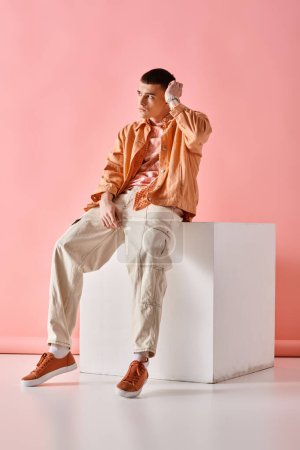 Foto de Fashionable man in beige shirt, pants and sneakers sitting on white cube on pink background - Imagen libre de derechos