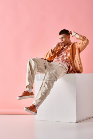 Foto de Joven en traje beige mirando hacia otro lado y sentado en cubo blanco sobre fondo rosa - Imagen libre de derechos