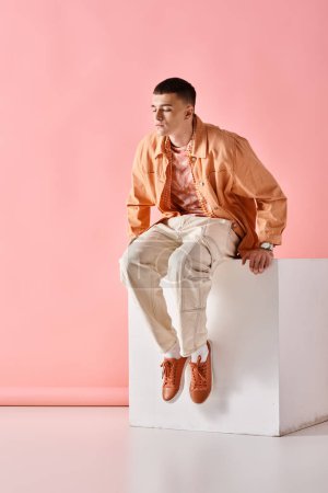 Téléchargez les photos : Homme à la mode en chemise beige, pantalon et baskets assis sur un cube blanc sur fond rose - en image libre de droit