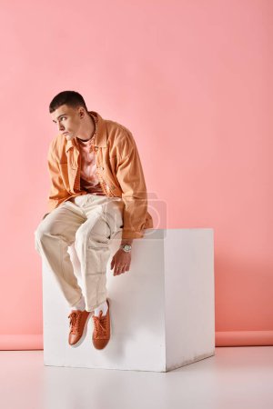Photo for Stylish man in beige outfit touching his hair and sitting on white cube on pink backdrop - Royalty Free Image