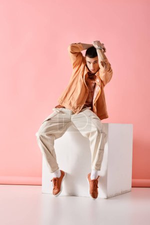 Téléchargez les photos : Homme à la mode avec les mains au-dessus de la tête et assis sur un cube blanc sur fond rose - en image libre de droit