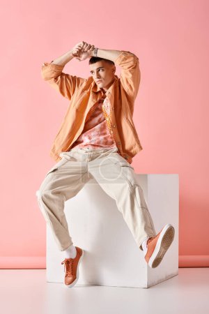 Fashionable young man in beige outfit touching his head and sitting on white cube on pink background