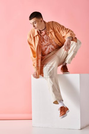 Photo for Stylish man in beige outfit looking down and sitting on white cube on pink background - Royalty Free Image