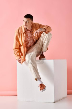 Homme à la mode en tenue tendance regardant vers le bas et assis sur un cube blanc sur fond rose