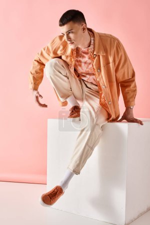 Full length image of stylish man in beige shirt, pants and boots on white cube on pink background