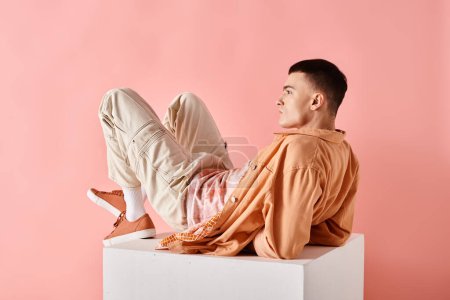Photo for Stylish man in peachy outfit touching his hair and lying on white cube on pink background - Royalty Free Image