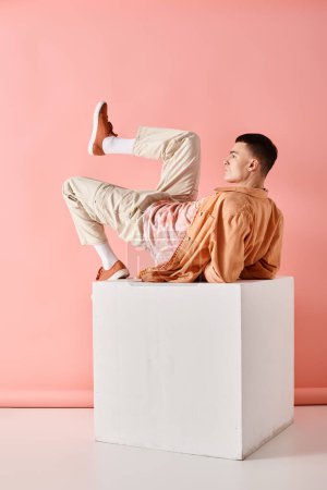 Photo for Side view photo of man in peach color outfit lying on white cube with leg up on pink background - Royalty Free Image