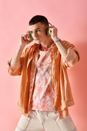 Stylish man in layered shirts with wireless headphones listening to music posing on pink backdrop