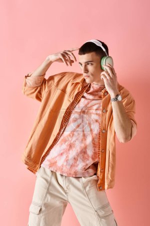 Fashionable man in layered outfit with wireless headphones dancing to music on pink backdrop