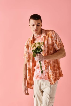 Téléchargez les photos : Prise de vue de mode de l'homme à la mode en tenue stratifiée tenant des fleurs sur fond rose - en image libre de droit