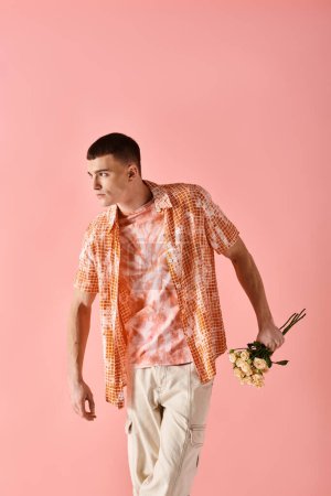 Portrait of stylish young man in layered outfit holding flowers posing on pink backdrop