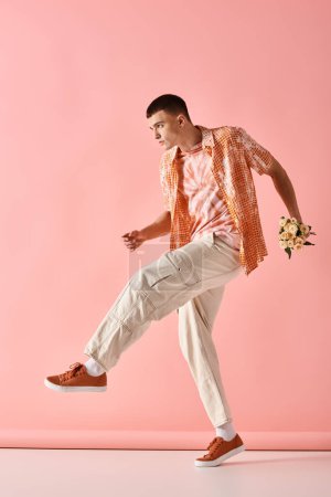 Téléchargez les photos : Image pleine longueur de l'homme à la mode en tenue stratifiée tenant des fleurs sur fond rose - en image libre de droit