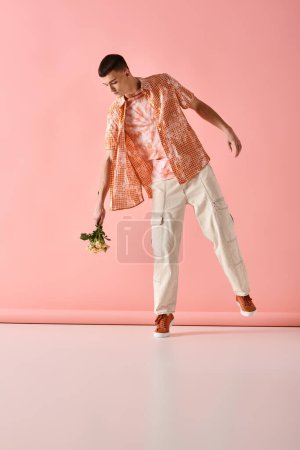 Téléchargez les photos : Image pleine longueur d'un bel homme en tenue de couleur pêche en couches tenant des fleurs sur fond rose - en image libre de droit