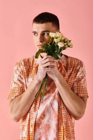 Téléchargez les photos : Bel homme en couches robe de couleur pêche tenant roses près du visage sur fond rose - en image libre de droit