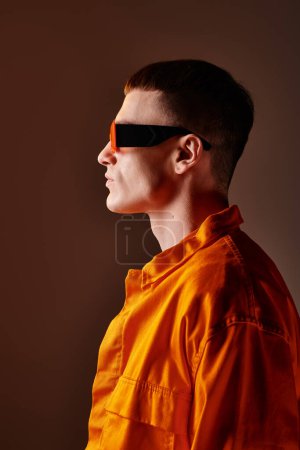 Foto de Retrato de moda vista lateral de hombre guapo en camisa naranja y gafas de sol sobre fondo marrón - Imagen libre de derechos