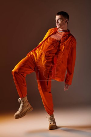 Foto de Moda tiro de longitud completa de hombre con estilo en mono naranja y chaqueta posando sobre fondo marrón - Imagen libre de derechos