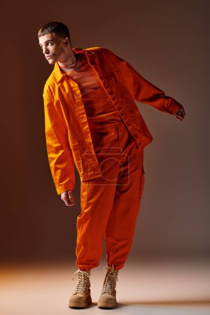 Ganztägiges Porträt eines modischen Mannes in orangefarbenem Overall und Jacke, der auf braunem Hintergrund posiert