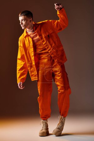 Foto de Tiro de longitud completa de hombre con estilo en mono naranja y chaqueta posando sobre fondo marrón - Imagen libre de derechos