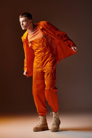 Foto de Tiro de longitud completa de hombre con estilo en mono naranja y chaqueta de pie sobre fondo marrón - Imagen libre de derechos