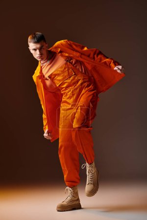 Foto de Moda tiro de longitud completa de hombre con estilo en mono naranja y chaqueta posando sobre fondo marrón - Imagen libre de derechos