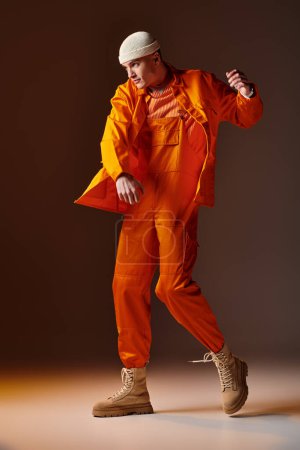 Foto de Tirador de cuerpo entero de hombre en mono naranja y chaqueta, gorro beige posando sobre fondo marrón - Imagen libre de derechos