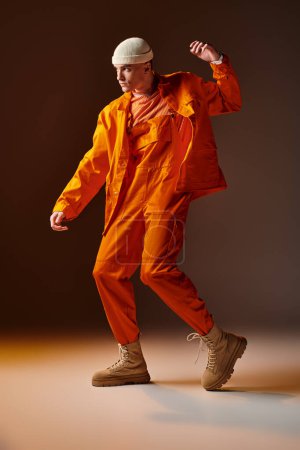 Foto de Joven hombre guapo en mono naranja y chaqueta, gorro beige posando sobre fondo marrón - Imagen libre de derechos